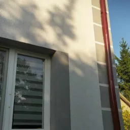 Fragment elewacji budynku po malowaniu w odcieniach szarości, z widoczną rynną w kolorze ceglastym i cieniem drzewa na ścianie.
