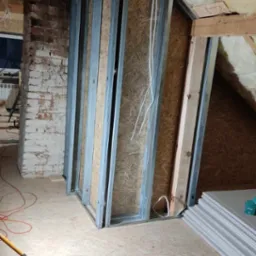 Konstrukcja ścianki działowej z metalowych profili i płyt OSB na poddaszu, widoczny fragment muru z cegły i składowane płyty gipsowo-kartonowe.