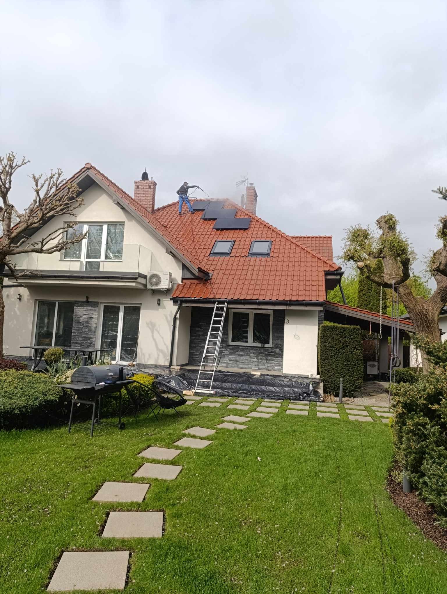 Pracownik w trakcie czyszczenia dachu z czerwonej dachówki myjką ciśnieniową; widoczne panele słoneczne, drabina oparta o dach, zabezpieczona folią elewacja domu.
