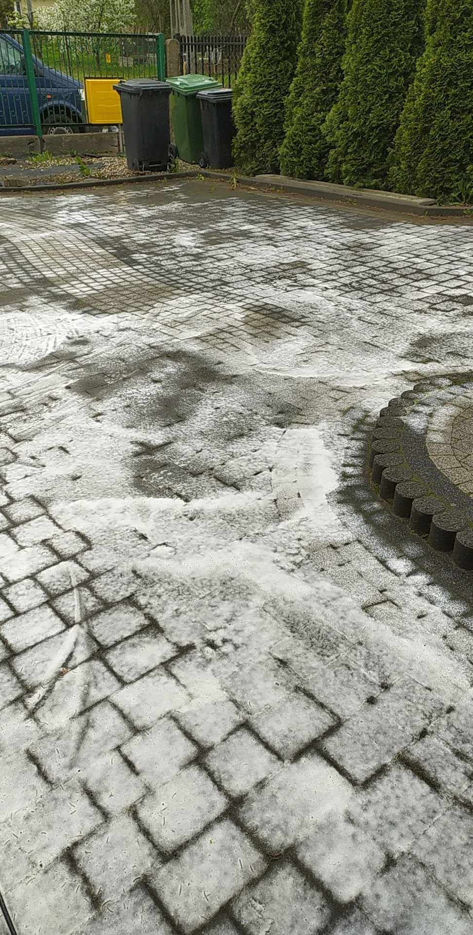 Mokra kostka brukowa po czyszczeniu, widoczne ślady wody i resztki zabrudzeń, w tle pojemniki na śmieci i krzewy.