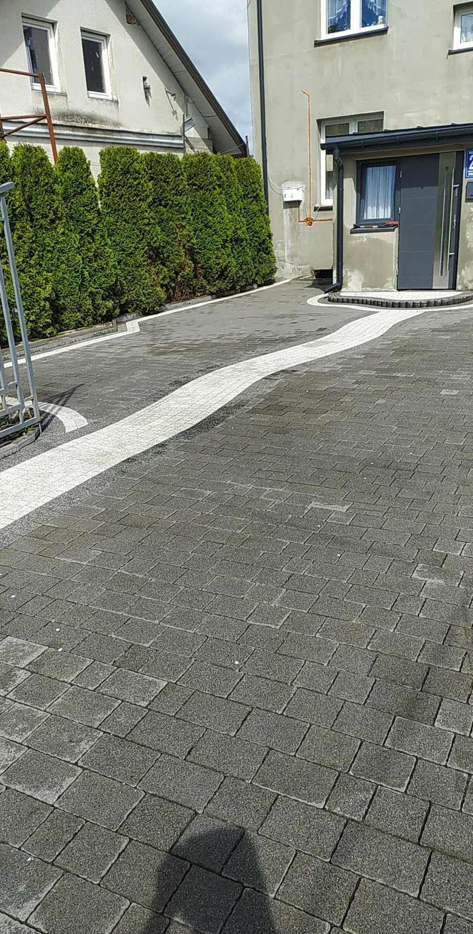 Szara kostka brukowa ułożona w łukowaty wzór z jasnymi elementami, otoczona zielonymi tujami i budynkiem z szarą elewacją.