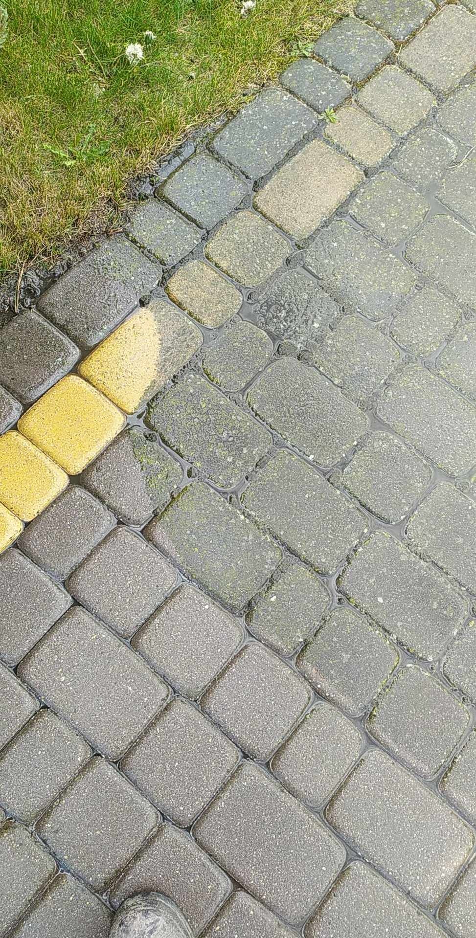 Mokre, szare kostki brukowe ułożone w jodełkę, z fragmentem żółtych kostek i zielenią w rogu. W zagłębieniach między kostkami stoi woda.