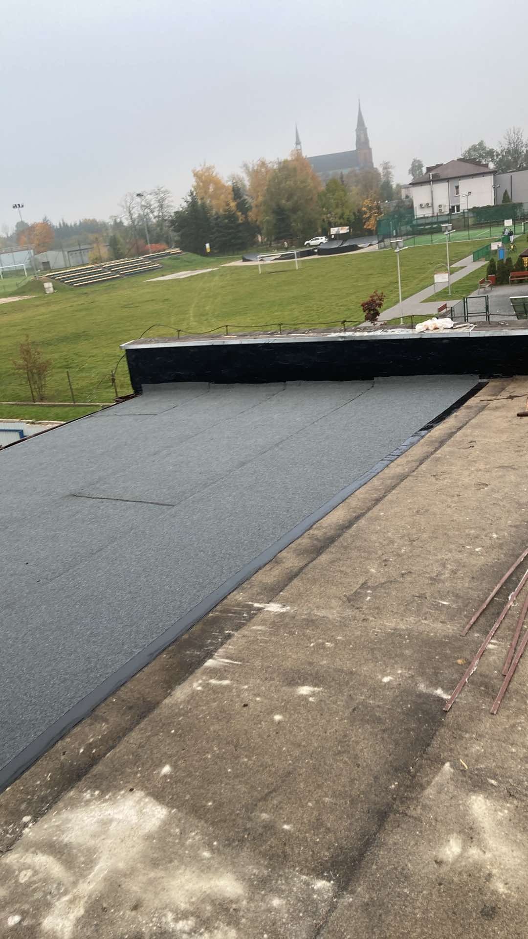 Płaski dach pokryty szarą papą termozgrzewalną, widoczne łączenia i krawędzie, w tle boisko sportowe i budynki w oddali, pochmurne niebo.
