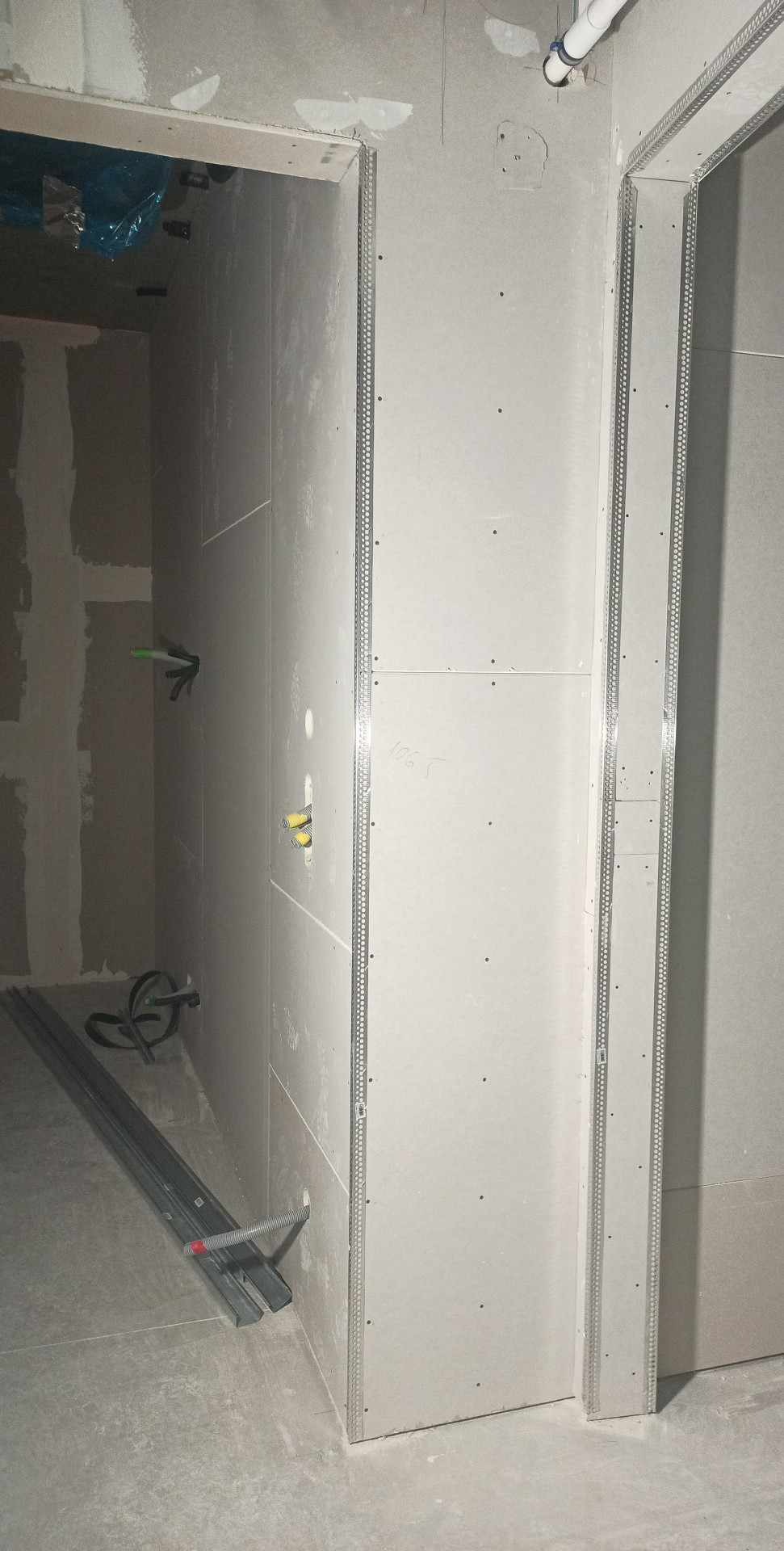 Wnętrze w trakcie prac wykończeniowych: ściany obłożone płytami gipsowo-kartonowymi, widoczne metalowe profile narożnikowe, instalacja elektryczna w ścianie oraz profile na podłodze.