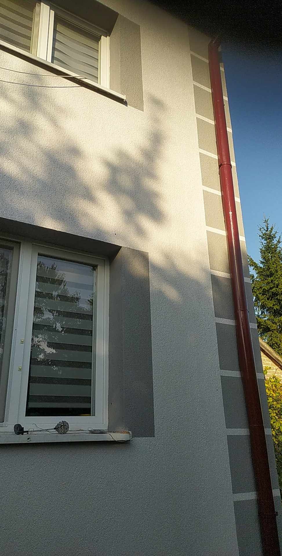 Fragment elewacji budynku po malowaniu w odcieniach szarości, z widoczną rynną w kolorze ceglastym i cieniem drzewa na ścianie.