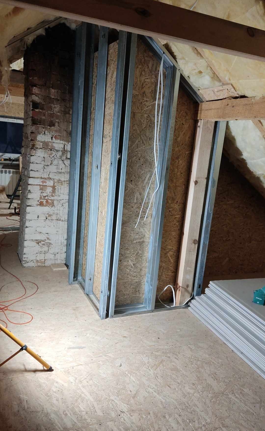 Konstrukcja ścianki działowej z metalowych profili i płyt OSB na poddaszu, widoczny fragment muru z cegły i składowane płyty gipsowo-kartonowe.