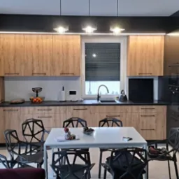 Nowoczesna kuchnia z drewnopodobnymi frontami szafek, czarnymi blatami i wysoką zabudową, uzupełniona o stół jadalny z czarnymi krzesłami o geometrycznym designie. Trzy lampy wiszące nad oknem.