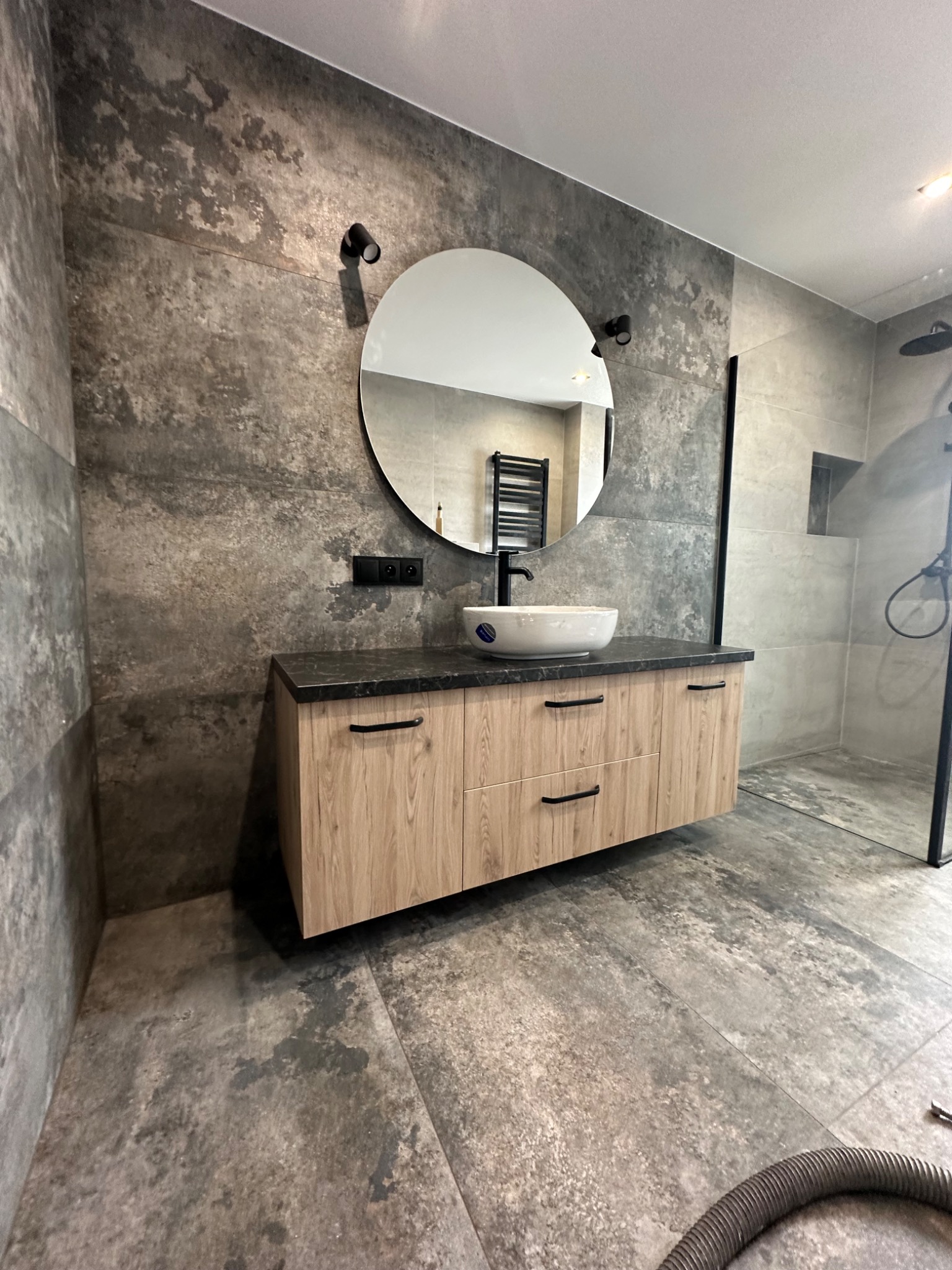 Nowoczesna łazienka z drewnopodobną szafką pod umywalkę, okrągłym lustrem, szarą mozaiką na ścianie i kabiną prysznicową typu walk-in, z widocznym wężem od odkurzacza na podłodze.