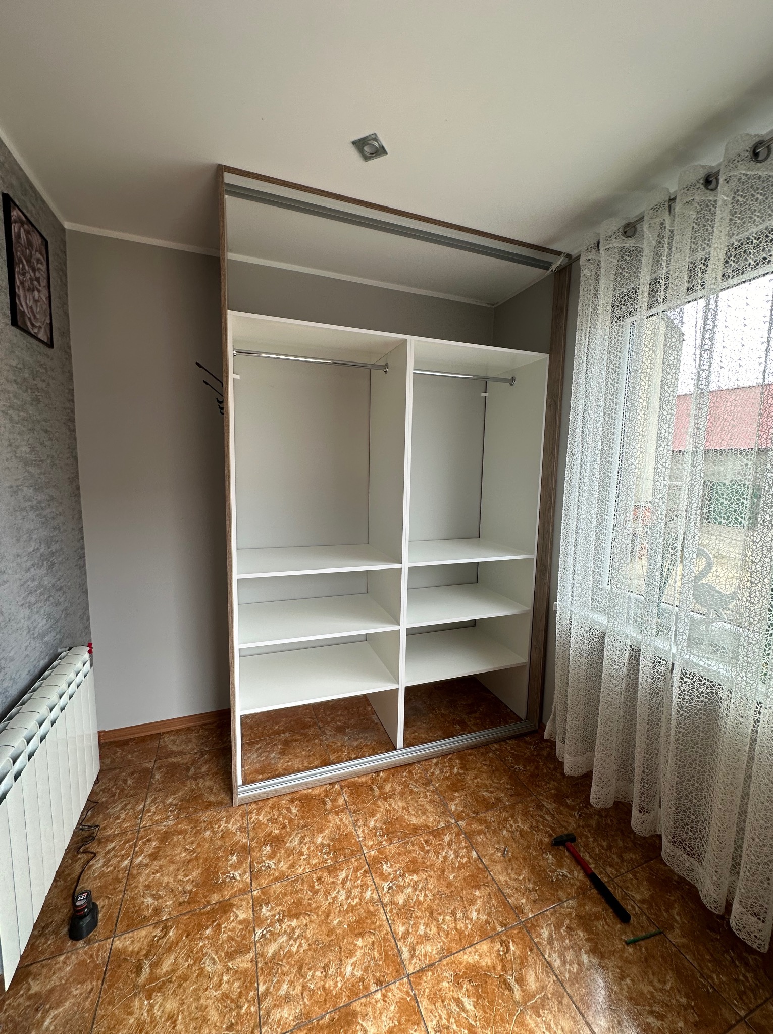 Wnętrze pokoju z częściowo zamontowaną białą szafą z przesuwnymi drzwiami, widoczne półki i drążki, na podłodze leży młotek i kołek, obok okna z ażurowymi zasłonami.