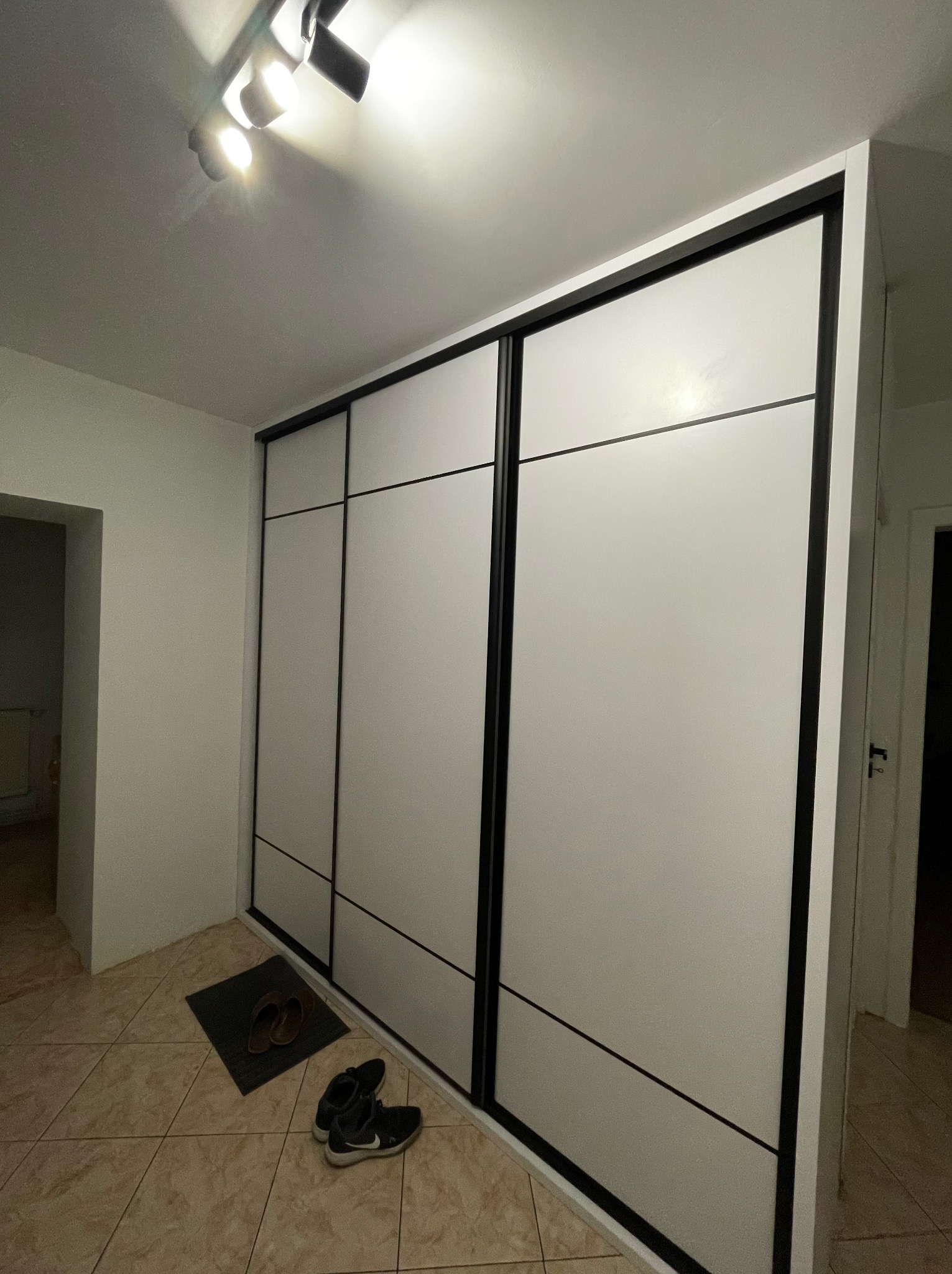 Biała szafa wnękowa z przesuwnymi drzwiami i czarnymi obramowaniami, widoczne buty i klapki na podłodze z terakoty, oświetlenie sufitowe.