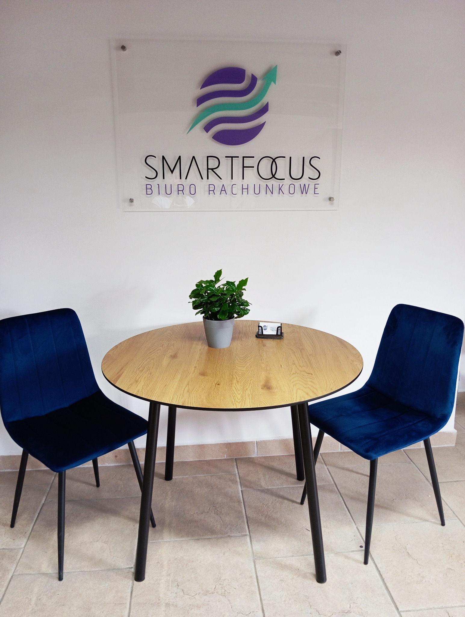 Wnętrze biura rachunkowego: okrągły stół z drewnianym blatem, dwa granatowe krzesła welurowe i roślina doniczkowa na tle białej ściany z logo firmy Smartfocus Biuro Rachunkowe.