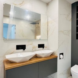 „DO-MEB” Dominik Sobczyk - Nowoczesna łazienka z dwiema białymi umywalkami o geometrycznym kształcie, osadzonymi na drewnianym blacie i szarych szafkach, z dużym lustrem na ścianie wyłożonej marmurowymi płytkami i widoczną...