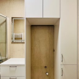 „DO-MEB” Dominik Sobczyk - Biała zabudowa meblowa w łazience z szafkami wiszącymi i umywalką z szufladami, widoczna kabina prysznicowa z lustrem i grzejnikiem drabinkowym w tle.