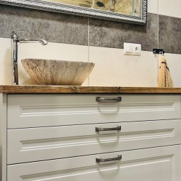 „DO-MEB” Dominik Sobczyk - Łazienkowa szafka z trzema szufladami w kolorze jasnoszarym, drewniany blat z umywalką w stylu kamiennej misy, chromowana bateria, lustro w ozdobnej ramie odbijające kabinę prysznicową.