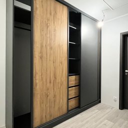 „DO-MEB” Dominik Sobczyk - Nowoczesna szafa przesuwna na wymiar z drewnianym frontem i czarnymi elementami, widok z boku, na podłodze panele imitujące drewno, jasne wnętrze z półkami i szufladami.