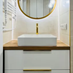 Nowoczesna łazienka z białą szafką pod umywalkę, złotym okrągłym lustrem z podświetleniem LED i złotą armaturą, na tle beżowych płytek.