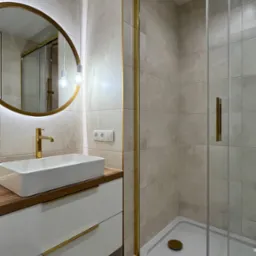 Nowoczesna łazienka z beżowymi płytkami, owalnym lustrem z podświetleniem LED, białą szafką z drewnianym blatem i złotymi uchwytami oraz kabiną prysznicową ze złotymi ramami.