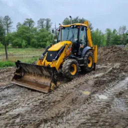 Żółta koparko-ładowarka JCB na błotnistym terenie z widocznymi śladami opon, w tle drzewa i pochmurne niebo.