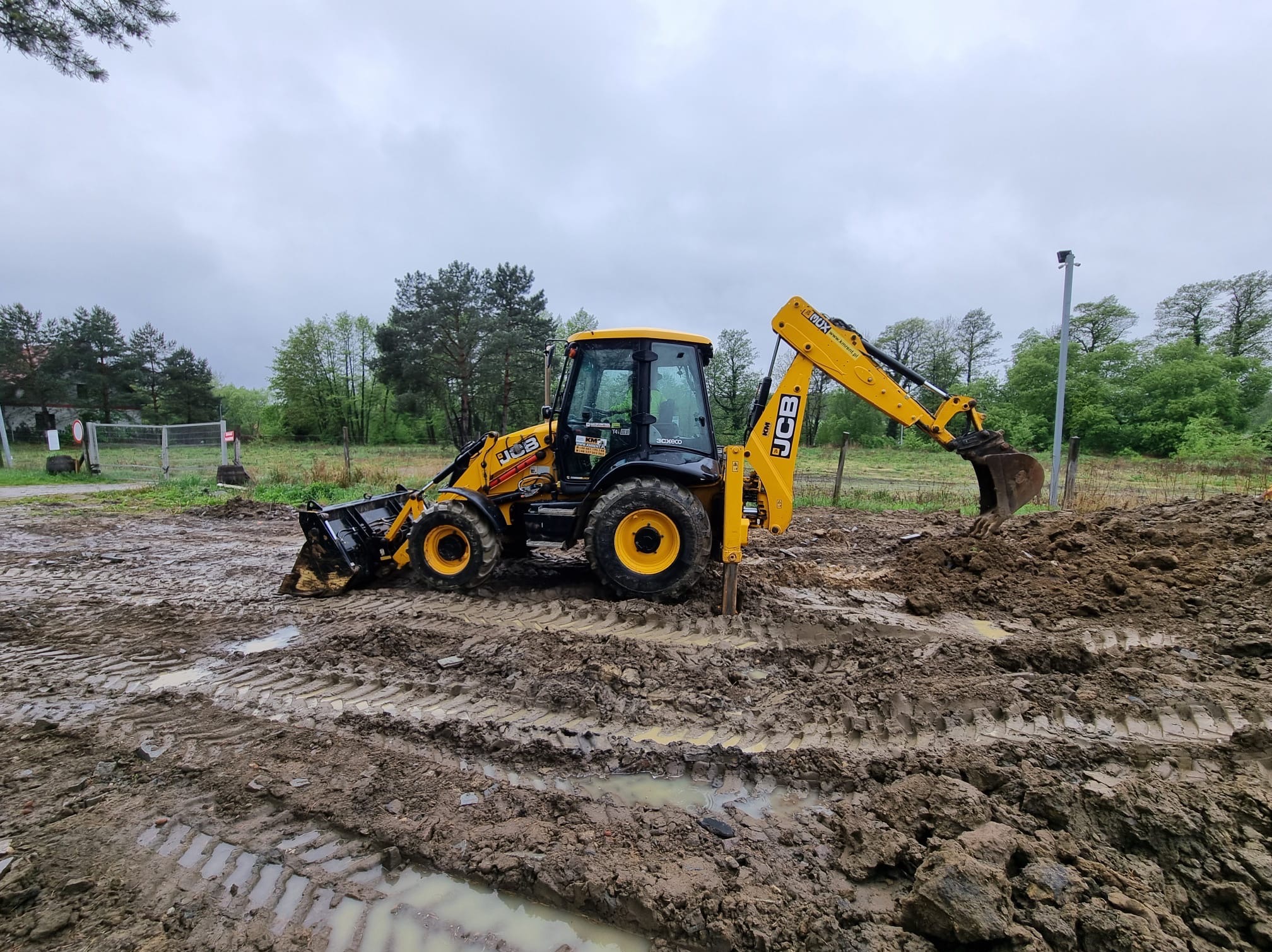 Żółta koparko-ładowarka JCB w błotnistym terenie podczas prac ziemnych, widoczne ślady opon i kopany grunt, pochmurne niebo w tle drzew