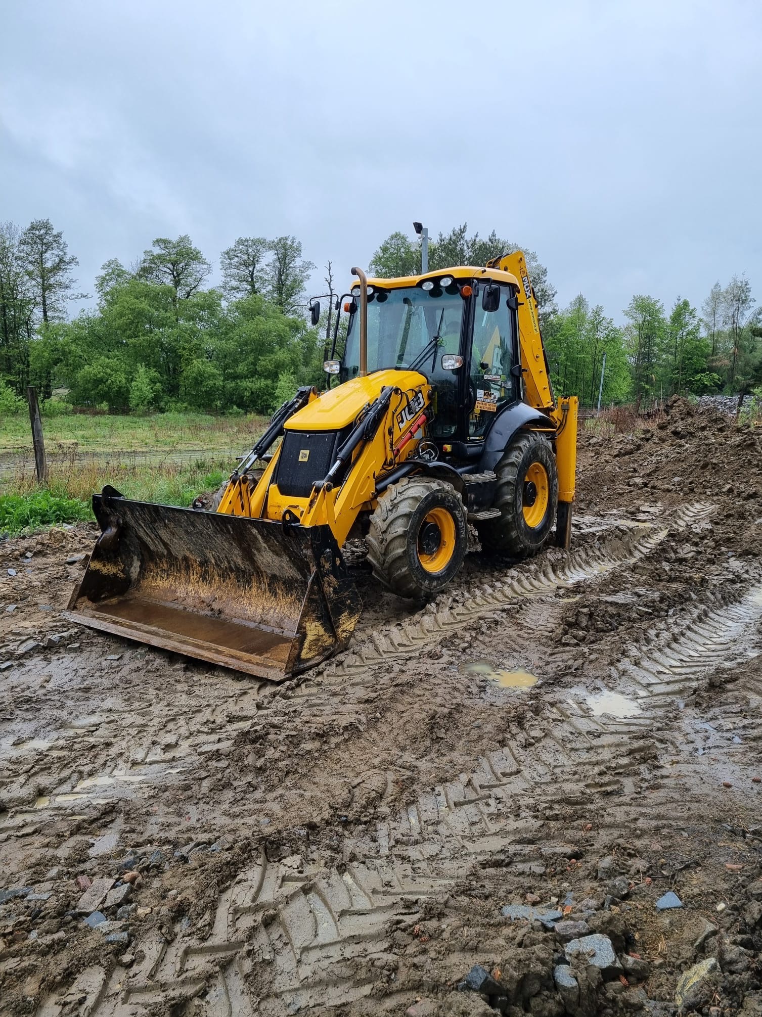 Żółta koparko-ładowarka JCB na błotnistym terenie z widocznymi śladami opon, w tle drzewa i pochmurne niebo.