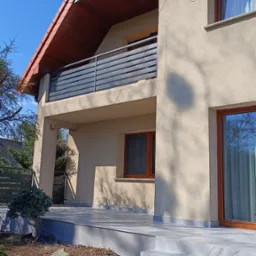 Nowoczesny dom z beżową elewacją, balkonem z poziomą, szarą balustradą i schodami wyłożonymi płytkami imitującymi marmur.