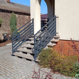 MT Metaltech - Betonowe schody wejściowe z szarą, metalową balustradą, prowadzące do domu z numerem 18 nad łukowym wejściem, otoczone kostką brukową i zielenią.