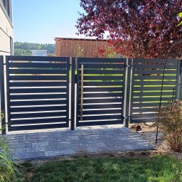 MT Metaltech - Nowoczesna, grafitowa brama wjazdowa z poziomymi panelami, otwierana na posesję wyłożoną szarą kostką brukową, z widokiem na drzewo o bordowych liściach i część budynku.