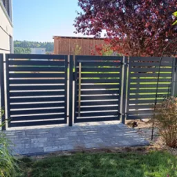 Nowoczesna, grafitowa brama wjazdowa z poziomymi panelami, otwierana na posesję wyłożoną szarą kostką brukową, z widokiem na drzewo o bordowych liściach i część budynku.