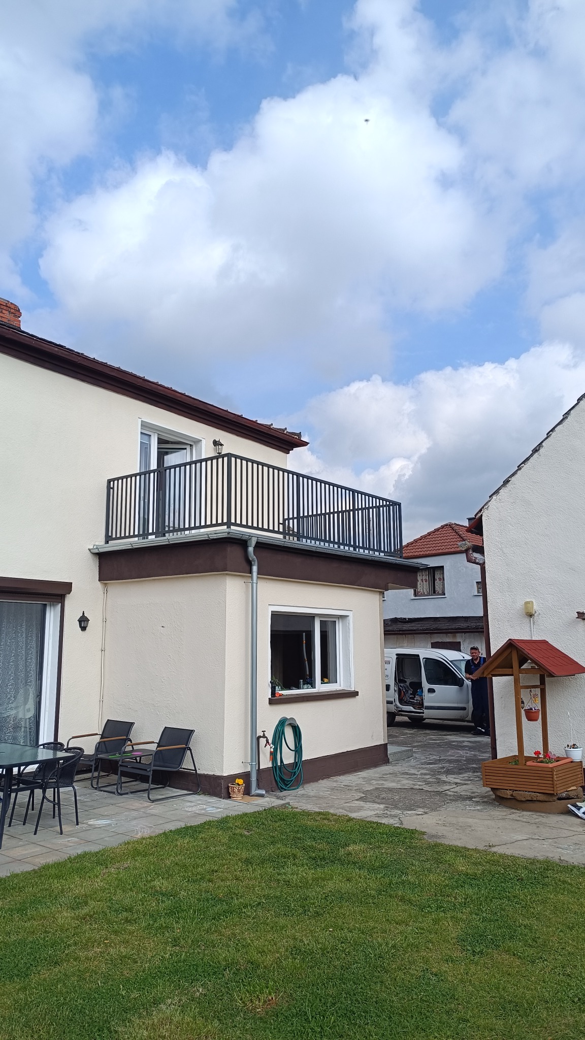 Balkon z czarną, metalową balustradą na jasnym budynku, widok z dołu, w tle biały samochód dostawczy i osoba.