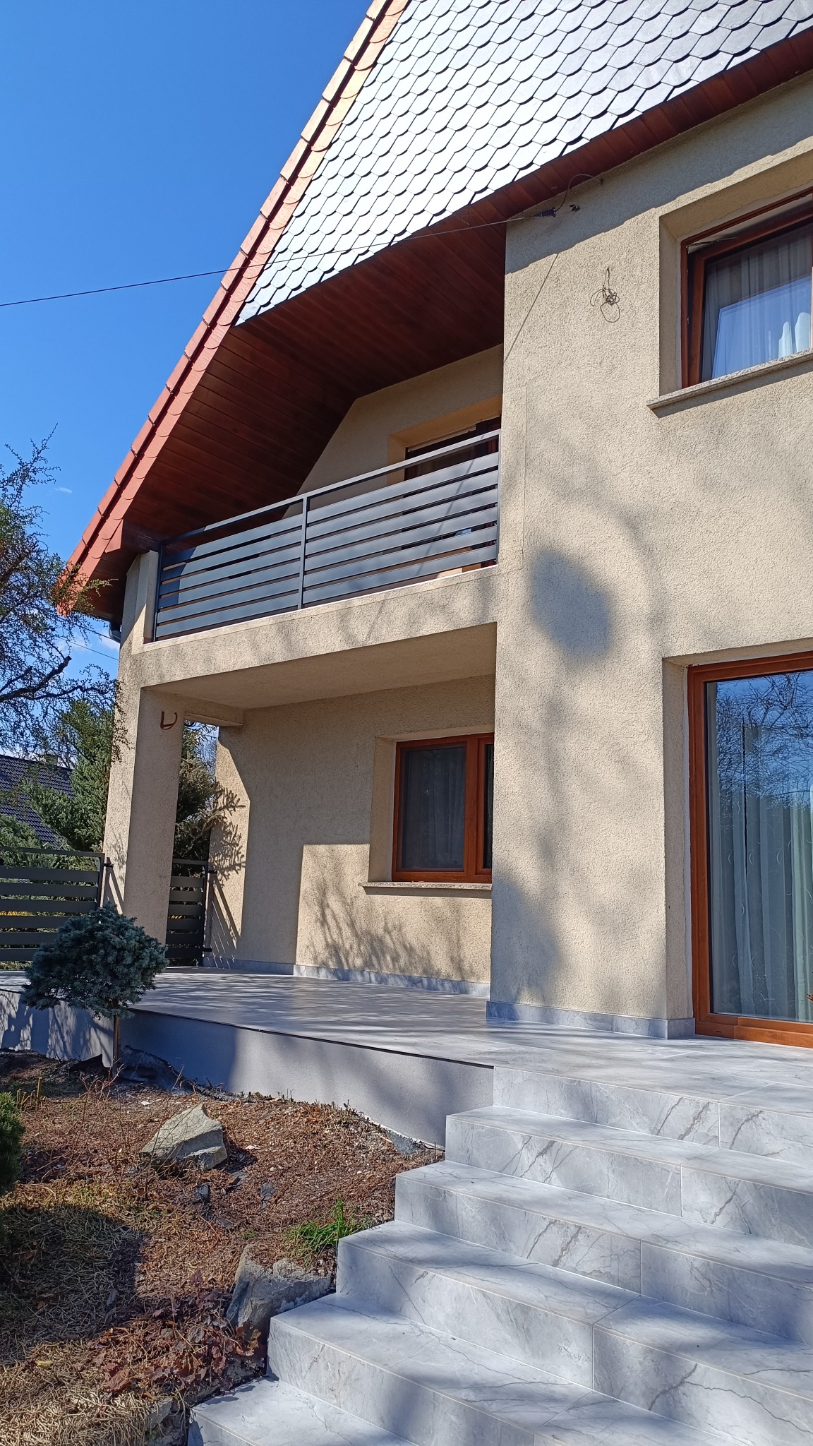 Nowoczesny dom z beżową elewacją, balkonem z poziomą, szarą balustradą i schodami wyłożonymi płytkami imitującymi marmur.