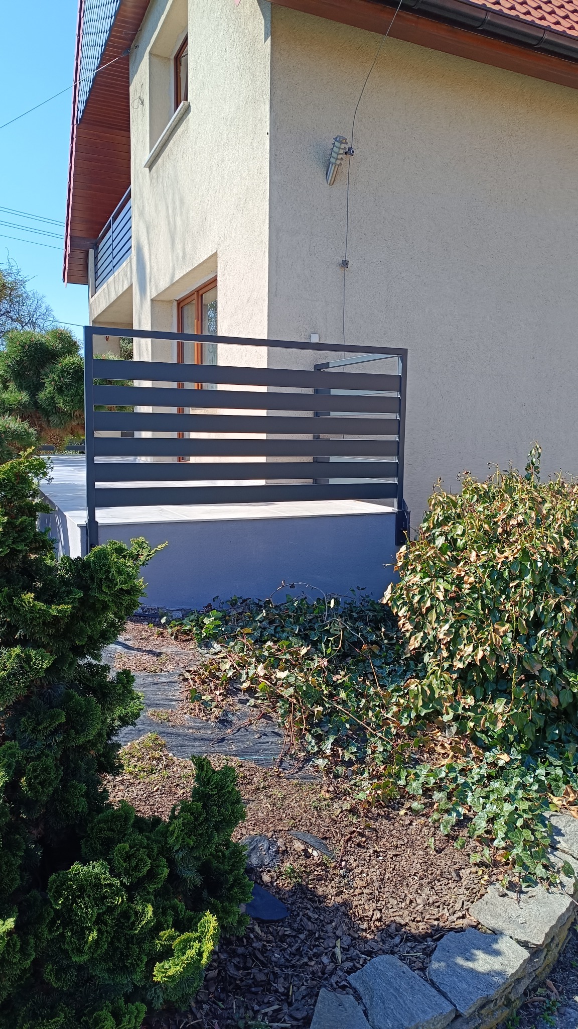 Nowoczesne, poziome ogrodzenie metalowe w kolorze antracytowym, zamontowane przy tarasie domu z kremową elewacją, widoczne rośliny ozdobne i fragment chodnika.