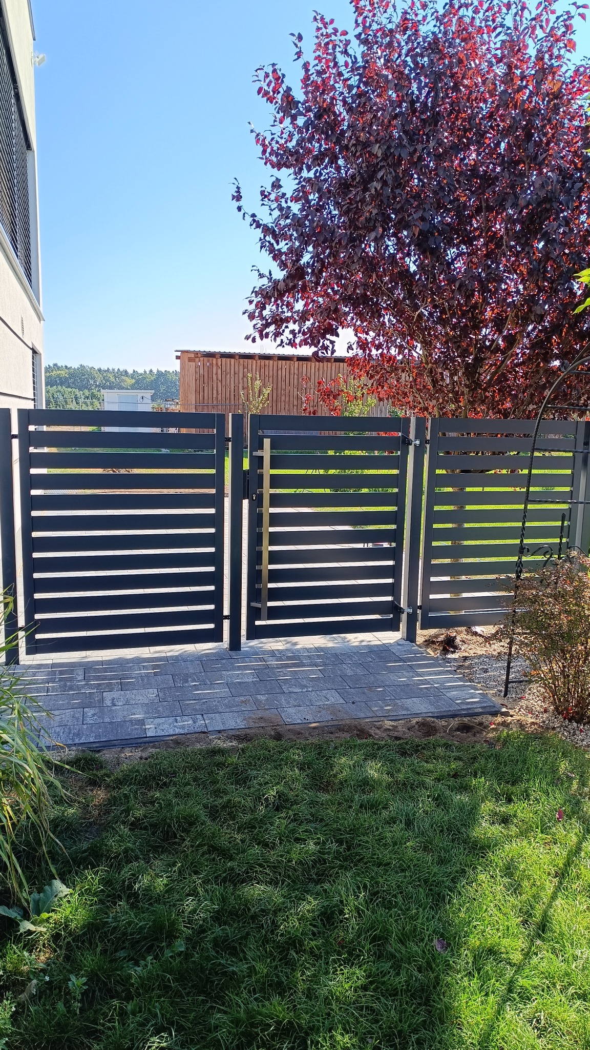 Nowoczesna, grafitowa brama wjazdowa z poziomymi panelami, otwierana na posesję wyłożoną szarą kostką brukową, z widokiem na drzewo o bordowych liściach i część budynku.