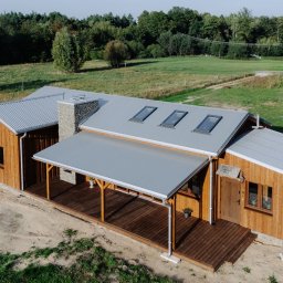 MBS Mateusz Marzec - Drewniany dom z szarą, falistą blachą na dachu i kamiennym kominem, widziany z lotu ptaka, otoczony zielenią. Widoczne okna dachowe i zadaszony taras.