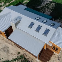 MBS Mateusz Marzec - Widok z góry na nowoczesny dom z drewnianą elewacją i szarym, falistym dachem z oknami dachowymi. Widoczny kamienny komin i drewniany taras.
