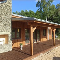 MBS Mateusz Marzec - Drewniany dom z zadaszonym tarasem i kamiennym kominkiem. Na tarasie stoi ozdobna donica z kwiatami. W tle widoczne drzewa.