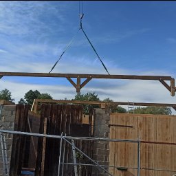 MBS Mateusz Marzec - Montaż drewnianej konstrukcji domu szkieletowego z użyciem dźwigu. Widoczny fragment ściany z desek, pracownik na rusztowaniu i budynek w stanie surowym w tle.