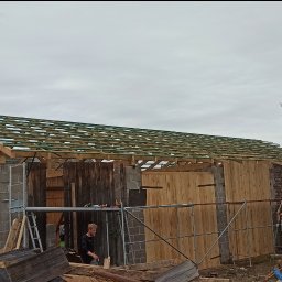 MBS Mateusz Marzec - Wznoszenie więźby dachowej na murowanym budynku z drewnianą elewacją. Widoczny pracownik i rozłożone narzędzia. W tle zielone drzewa.