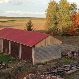 MBS Mateusz Marzec - Budynek gospodarczy z czerwonym dachem na tle pól uprawnych i drzew. Elementy konstrukcyjne z cegły, betonu i drewna. Widoczne pozostałości materiałów budowlanych.