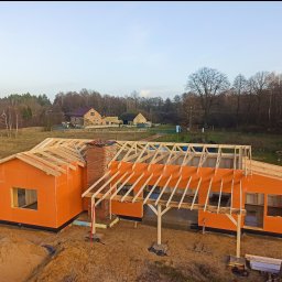 MBS Mateusz Marzec - Budowa domu w stanie surowym otwartym: pomarańczowe płyty elewacyjne, drewniana konstrukcja dachu, komin z cegły. W tle zabudowa wiejska i las.