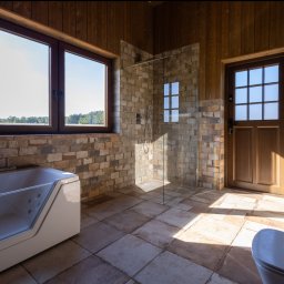 MBS Mateusz Marzec - Rustykalna łazienka z kabiną prysznicową, wanną z hydromasażem i drewnianymi elementami wykończenia. Widok na jezioro przez okno.