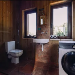 Rustykalna łazienka z drewnianymi ścianami i ceglaną zabudową umywalki. Widoczna toaleta, kabina prysznicowa i pralka Electrolux.