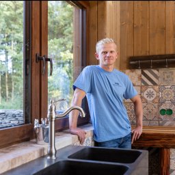 MBS Mateusz Marzec - Wnętrze domu z drewnianymi ścianami i oknem z widokiem na zieleń. Mężczyzna w niebieskiej koszulce opiera się o blat przy zlewie, z tyłu ściana z dekoracyjnymi płytkami.