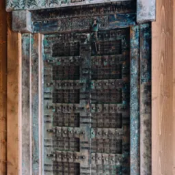 Rustykalne, dwuskrzydłowe drzwi z dekoracyjnym nadprożem i metalowymi okuciami, zabezpieczone kłódką, umieszczone w drewnianej ścianie z desek.