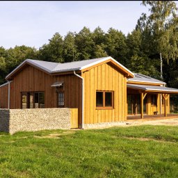 MBS Mateusz Marzec - Drewniany dom z szarym dachem i kamiennym ogrodzeniem na tle zieleni. Widoczne okna, drzwi i zadaszony taras. Jasne drewno elewacji, naturalne światło.