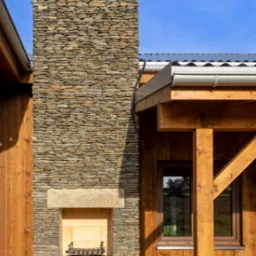 Kamienny komin zewnętrzny z grillem, przylegający do drewnianej konstrukcji z oknem. Widoczne elementy dachu i niebieskie niebo w tle.