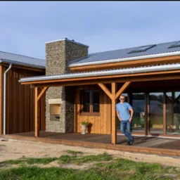 Drewniany dom z kamiennym kominem i zadaszonym tarasem, na którym stoi mężczyzna. Dach pokryty blachą, okna z brązowymi ramami. Całość w naturalnym otoczeniu.