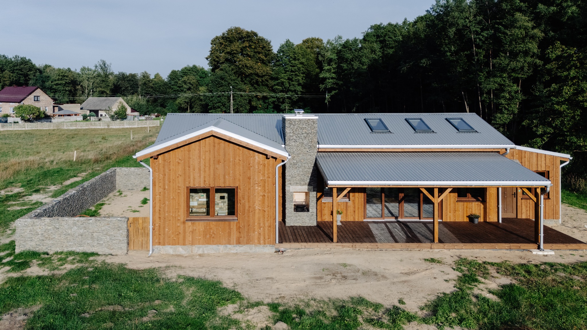 Nowoczesny dom z drewna z szarym dachem z blachy trapezowej i kamiennym kominem, otoczony zielenią. Widoczne okna dachowe i drewniany taras.