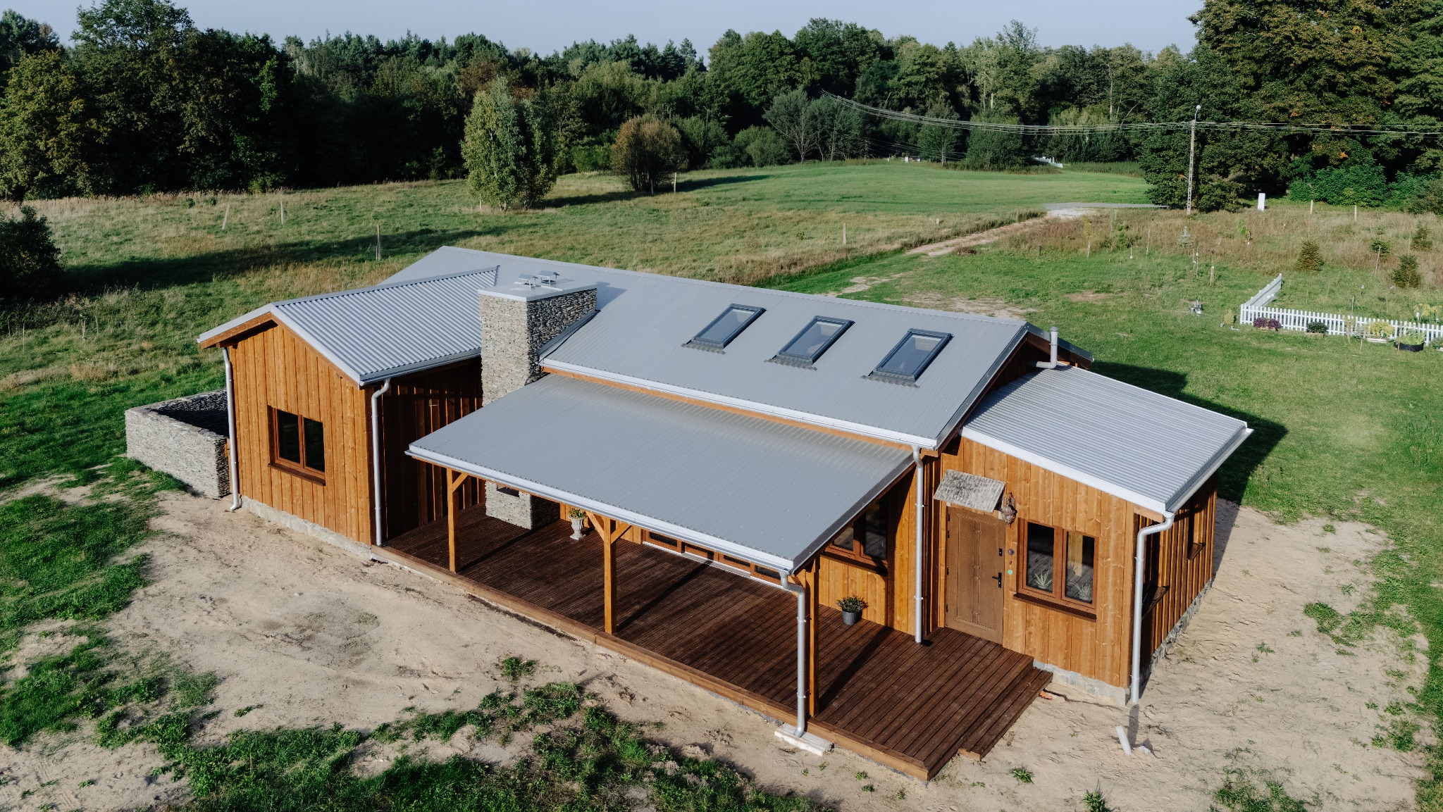 Drewniany dom z szarą, falistą blachą na dachu i kamiennym kominem, widziany z lotu ptaka, otoczony zielenią. Widoczne okna dachowe i zadaszony taras.