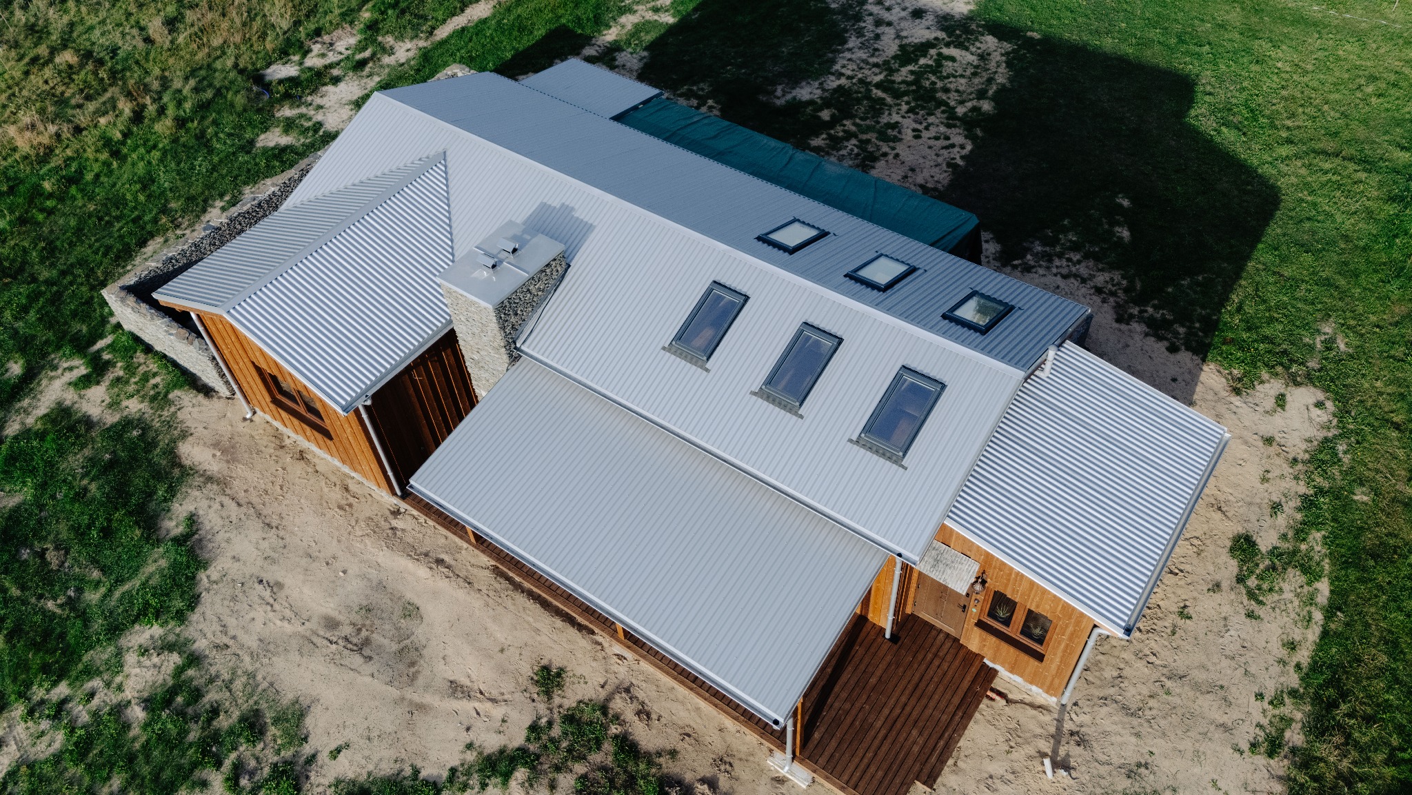 Widok z góry na nowoczesny dom z drewnianą elewacją i szarym, falistym dachem z oknami dachowymi. Widoczny kamienny komin i drewniany taras.