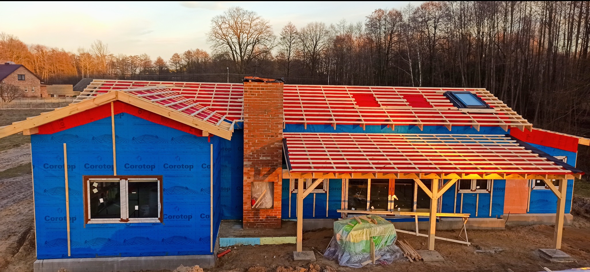 Budowa domu szkieletowego w trakcie: widoczna drewniana konstrukcja dachu z oknem, komin z cegły, ściany zabezpieczone folią Corotop, widok z góry na tle lasu.